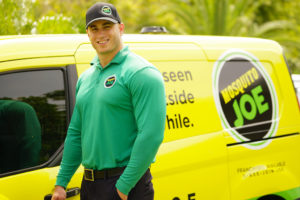 A smiling Mosquito Joe technician wearing a black hat and green long sleeve is posing for a picture next to a yellow service van.